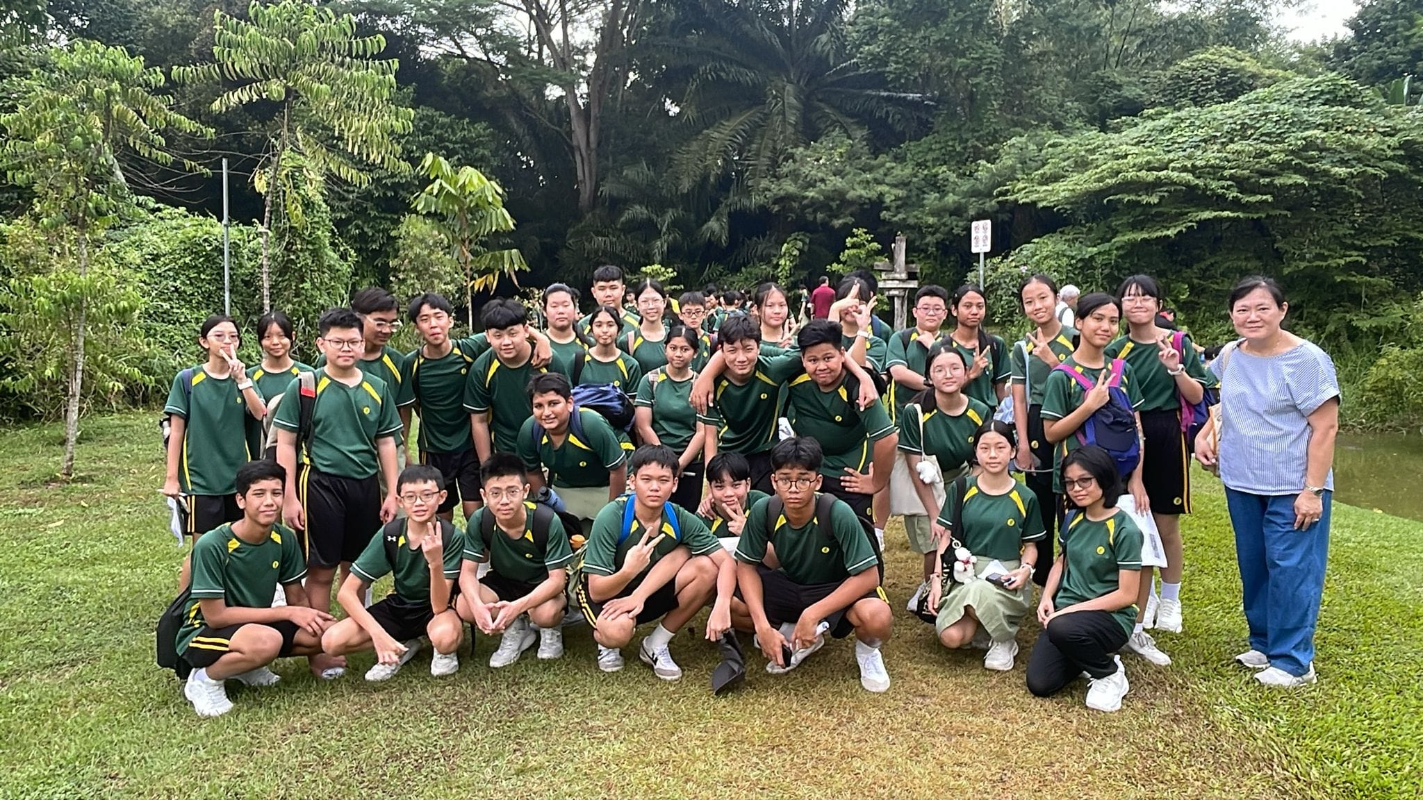 Photograph of a large group of students and two adults posing outdoors on grass near trees and a small body of water. Most students wear matching green and yellow sports uniforms, while one adult wears a light blue shirt and blue pants, indicating a school or team outing.